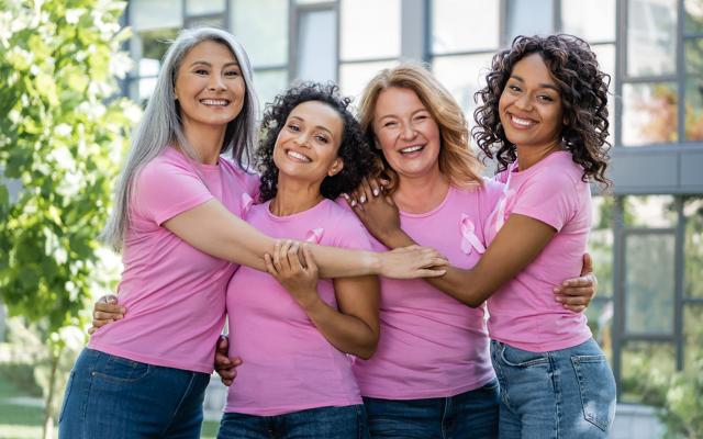 Four people wearing pink t-shirts and pink ribbon pins stand together outdoors, smiling and embracing to show support for breast cancer awareness.